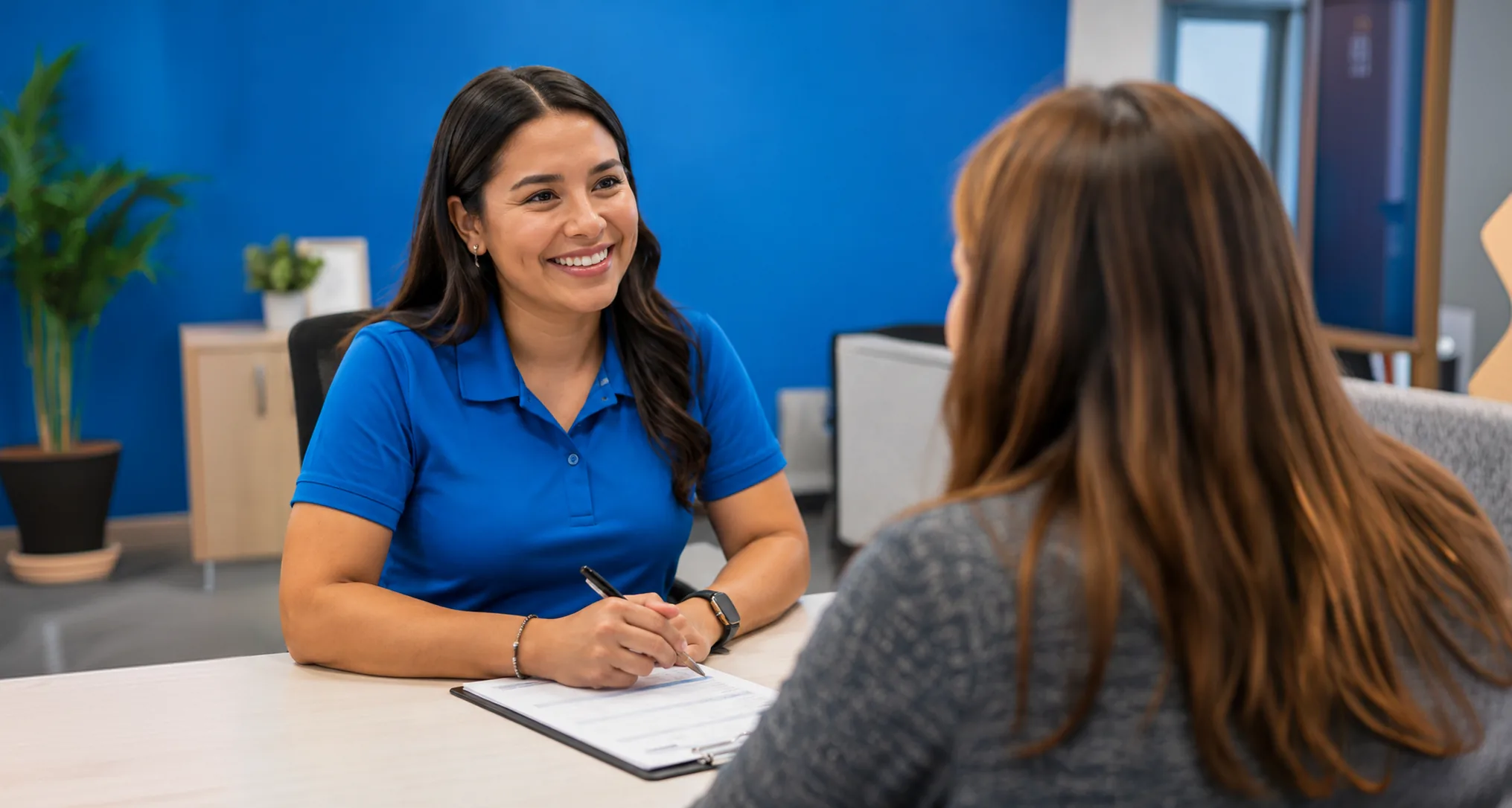 A smiling agent in a blue polo shirt fills in a form at a desk across from a client.