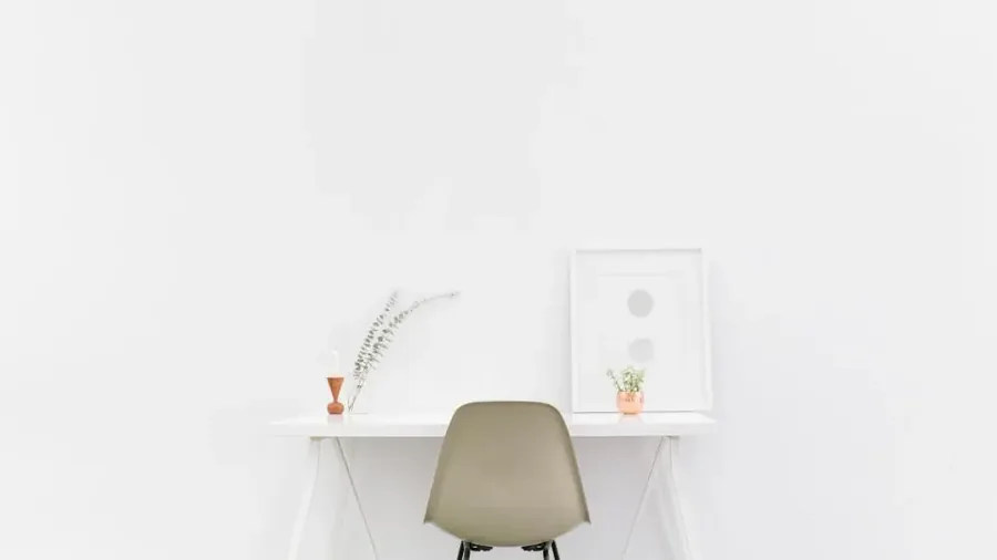 A tidy white desk with an empty light-colored chair tucked beneath it, a framed print and a small plant in a vase, in a bare, brightly lit home office with no one at work