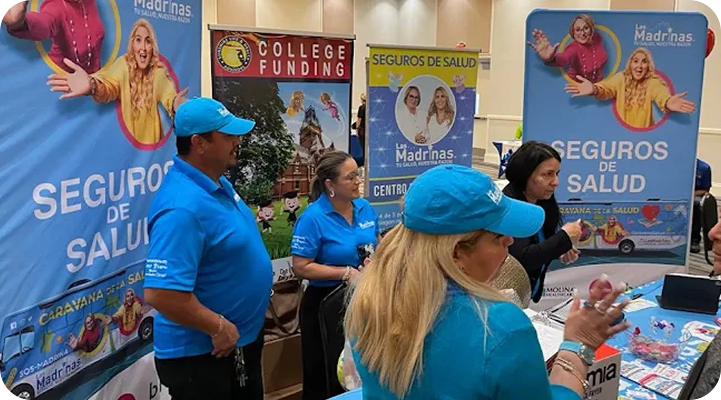 Integrantes del equipo de Madrinas con polos y gorras celestes conversan con visitantes en la mesa de un evento comunitario, frente a pancartas de Seguros de Salud y College Funding con las madrinas ilustradas