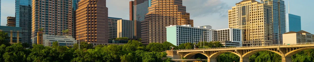 El horizonte del centro de Austin en un día despejado, con torres de vidrio y piedra de distintas alturas sobre un puente en arco y una línea de árboles verdes junto al agua