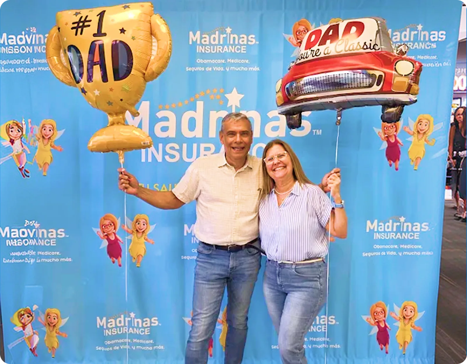 En un evento comunitario de Madrinas, un hombre con camisa color crema y una mujer con blusa a rayas sonríen frente a un fondo azul de Madrinas Insurance, con un globo dorado de trofeo de número uno para papá y un globo rojo de auto clásico