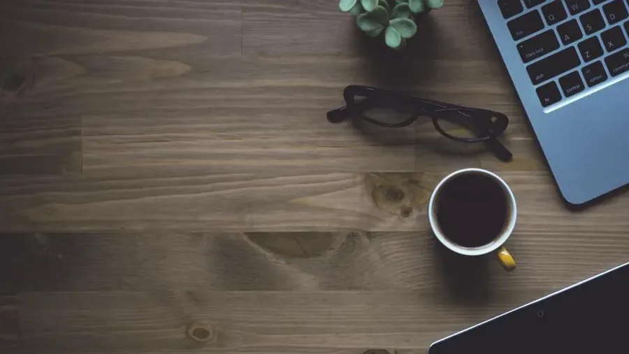 Overhead view of a dark wooden desk with the corner of an open laptop, a cup of black coffee, a pair of reading glasses and a tablet, in the warm light of a home workspace