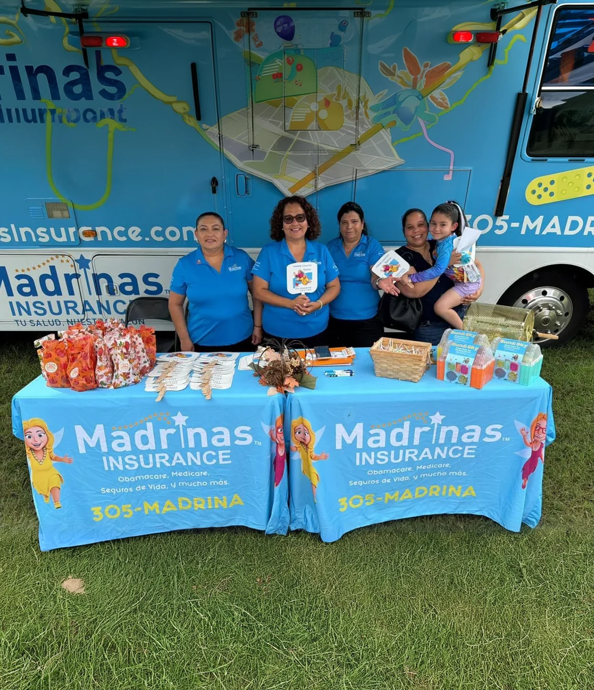 Three women in light-blue Madrinas Insurance polo shirts and a woman holding a young girl stand behind an outdoor table draped in Madrinas tablecloths printed with 305-MADRINA, set with gift bags, paper fans and toys, a Madrinas mobile unit parked behind them on the grass.